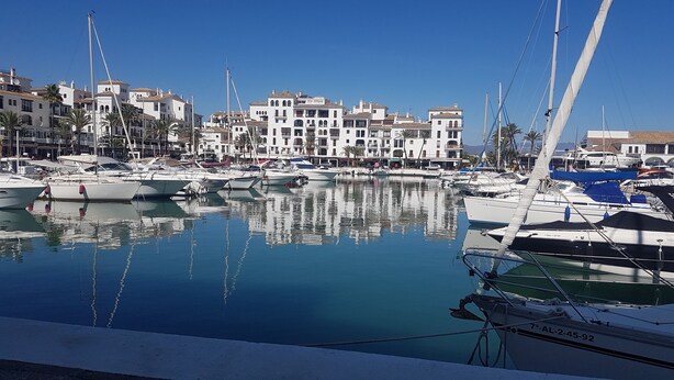 Un puerto deportivo con barcos amarrados, rodeado de edificios de varios pisos con balcones y un cielo azul despejado.
