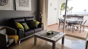 Una sala de estar con sofá, sillón y mesa de centro. Tiene un balcón con mesa y sillas.