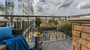 Een balkon met een rieten stoel, een metalen tafel met fruit en een glas wijn, en een uitzicht op gebouwen en een straat eronder.