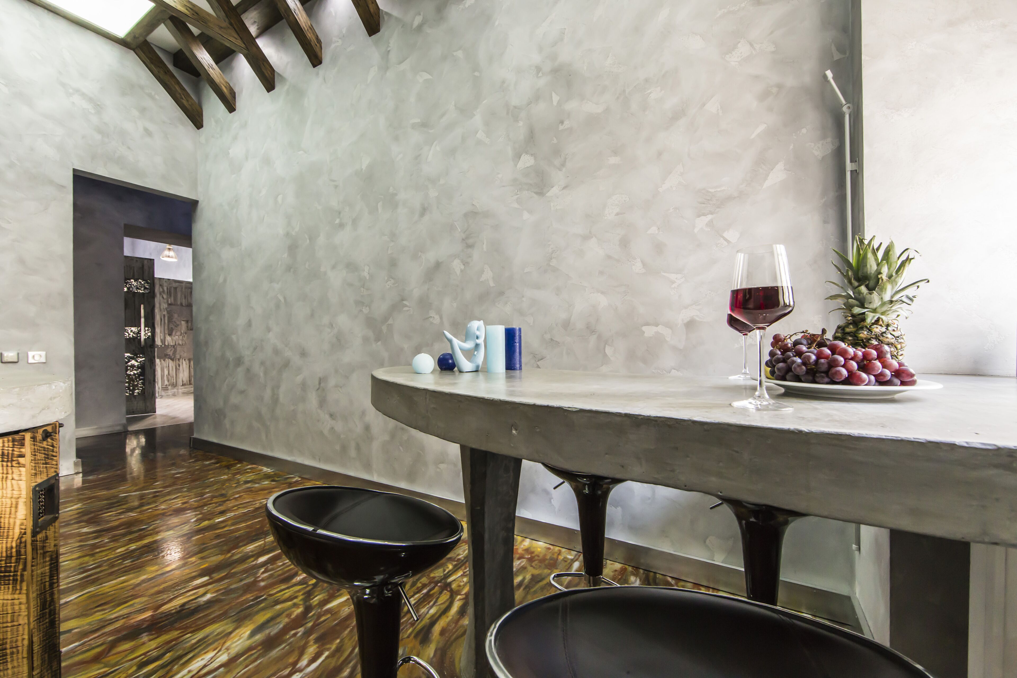 A modern dining area with a round table, two black chairs, a glass of red wine, a pineapple, and a plate of grapes.