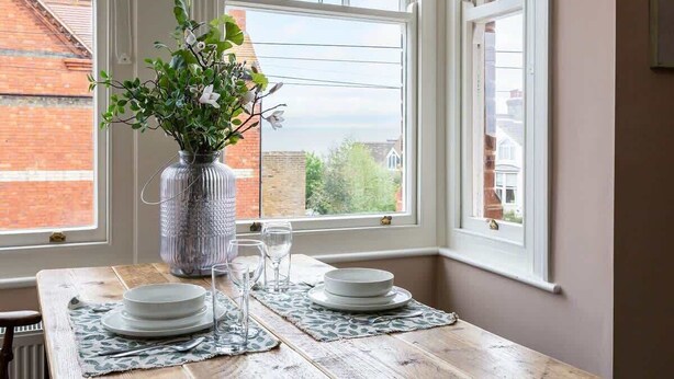 Una mesa de comedor preparada con platos, vasos y un florero con flores, ubicada junto a una ventana con vista a un edificio de ladrillo.