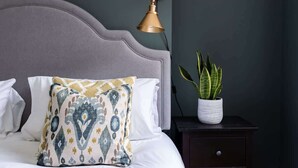 Una cama con almohadas estampadas, una lámpara de noche, una mesita de noche oscura y una planta en maceta.