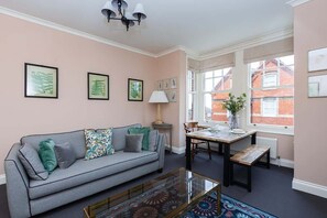 Ein Zimmer mit grauem Sofa, einem Esstisch mit Stühlen, einer Lampe und gerahmten Pflanzenbildern an der Wand.