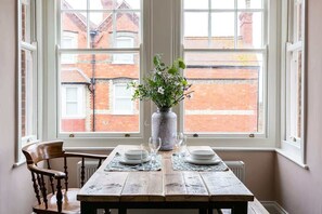Una mesa de comedor con platos y vasos, un florero con flores y una silla en una habitación con ventanas grandes.