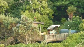 A wooden deck with a covered seating area, surrounded by dense greenery and trees. - 4~Short walk~Beach~Pubs~Shops: Chlorine safe Pool and Hot tub, Games room, WiFi  (Portreath)