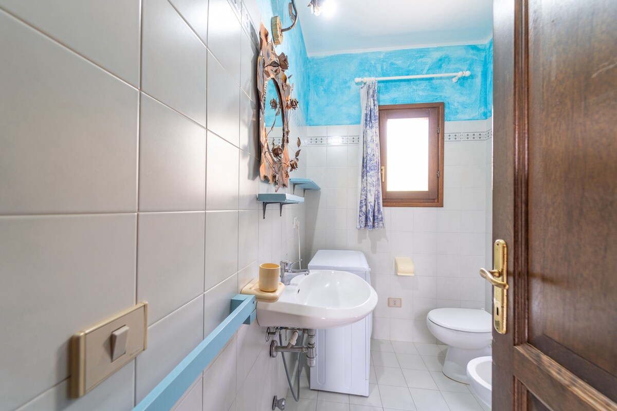 A bathroom with white tiles, a sink, a toilet, and a window with a blue curtain.