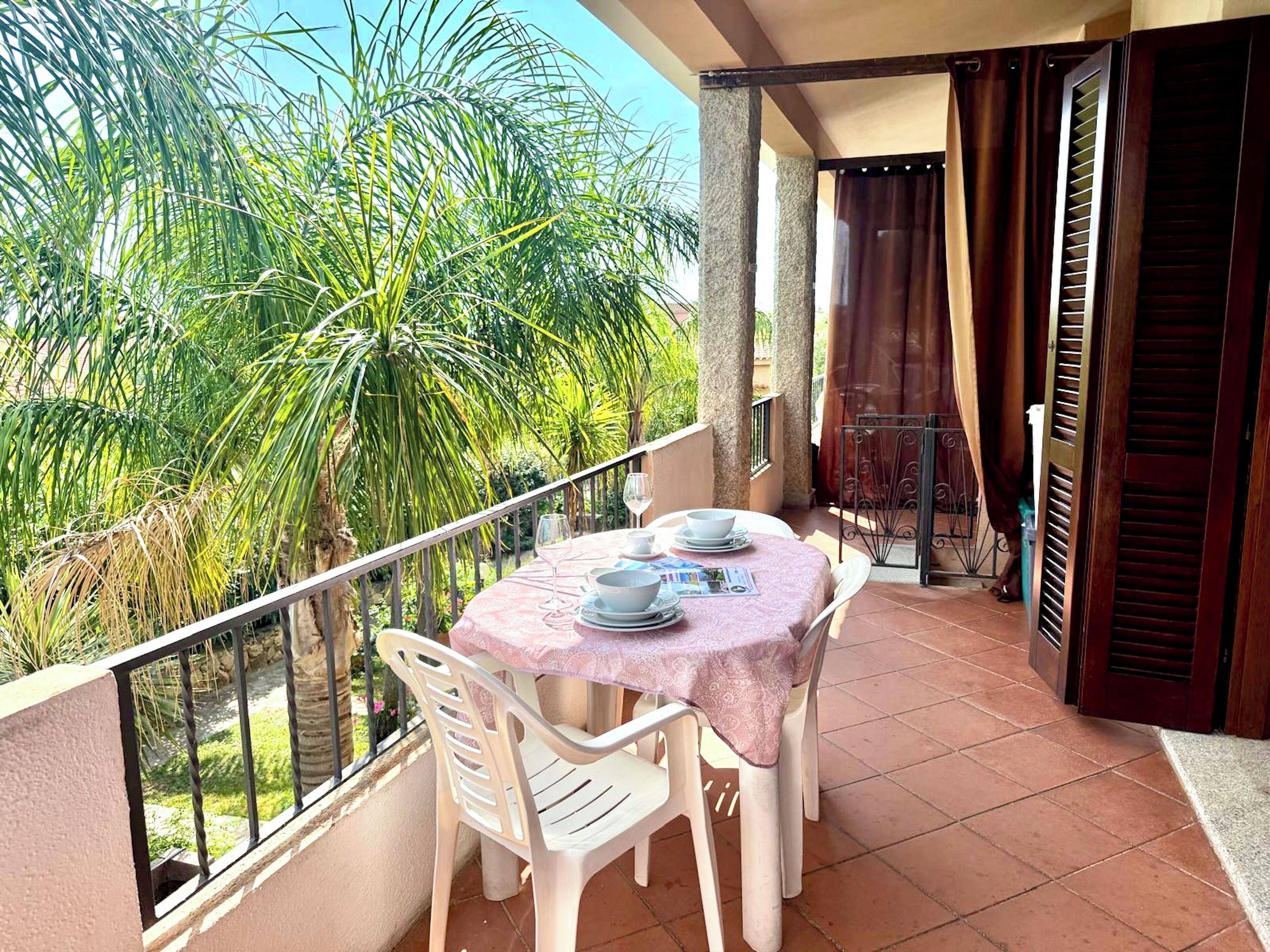 A balcony with a table set for two, surrounded by chairs, overlooking a garden with palm trees.
