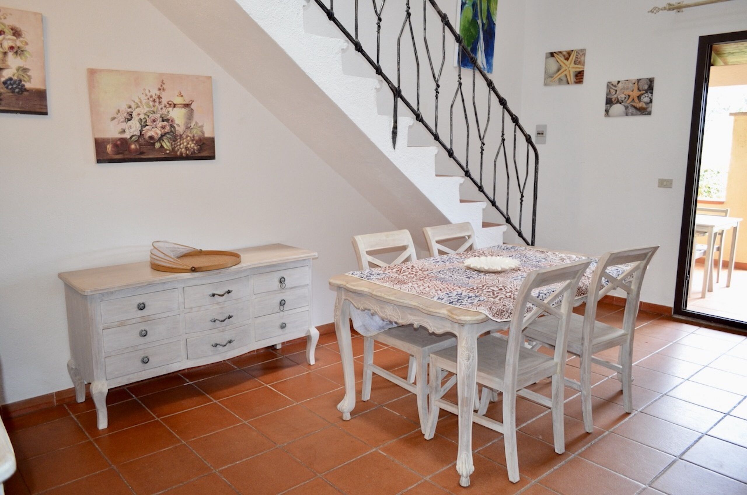 A dining area with a white table and chairs, a white cabinet with drawers, and a staircase with a black railing.