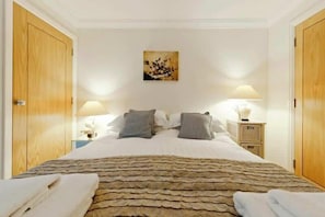 A bedroom with a stone-patterned bedspread, two bedside tables with lamps, a painting on the wall, and wooden doors.