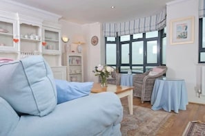 A living room with a light blue sofa, a wooden coffee table, a wicker chair, a small round table, and a large window with curtains.
