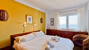 Una habitación de hotel con cama, ventana con cortinas, una silla de mimbre y decoración en la pared.
