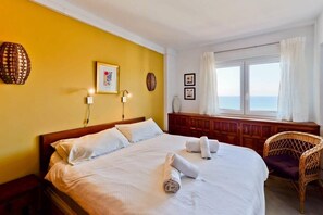 Une chambre d’hôtel avec un lit, une fenêtre avec des rideaux, un fauteuil en osier et des éléments de décoration murale.