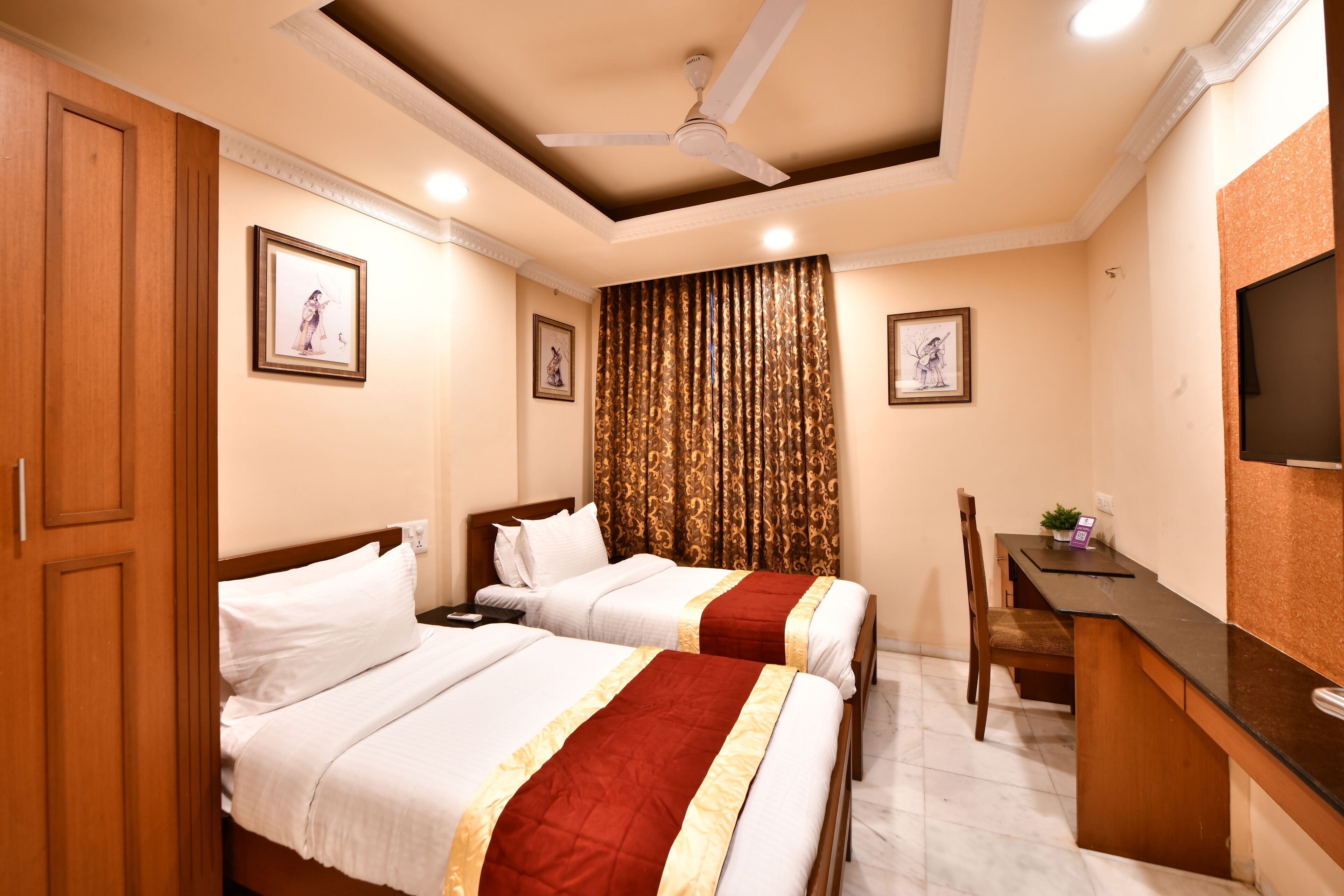 A hotel room with two beds, a desk, a chair, a ceiling fan, and a TV.