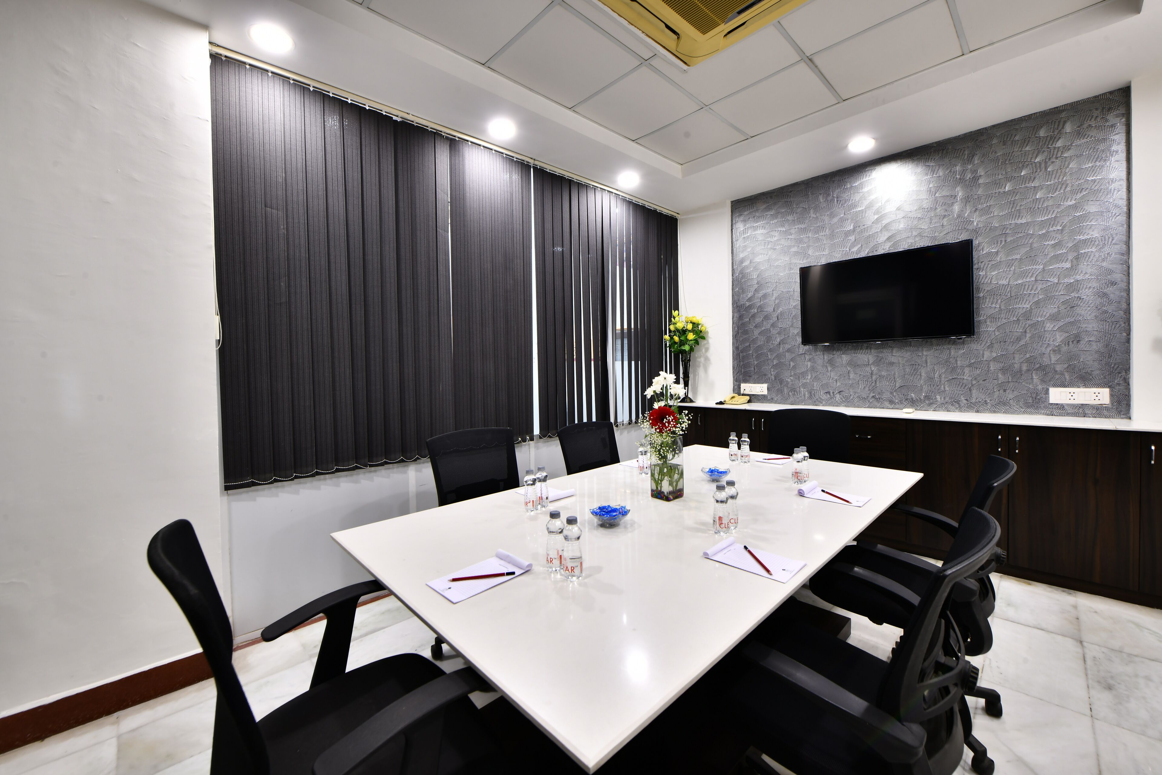 A conference room with a white table, black chairs, a TV on the wall, and a flower vase.