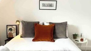 Un lit bien fait, avec du linge de lit blanc et un plaid gris et moelleux. Il y a deux oreillers sur le lit, un orange et un gris. Une petite table avec une lampe et un tableau encadré se trouve à côté du lit.