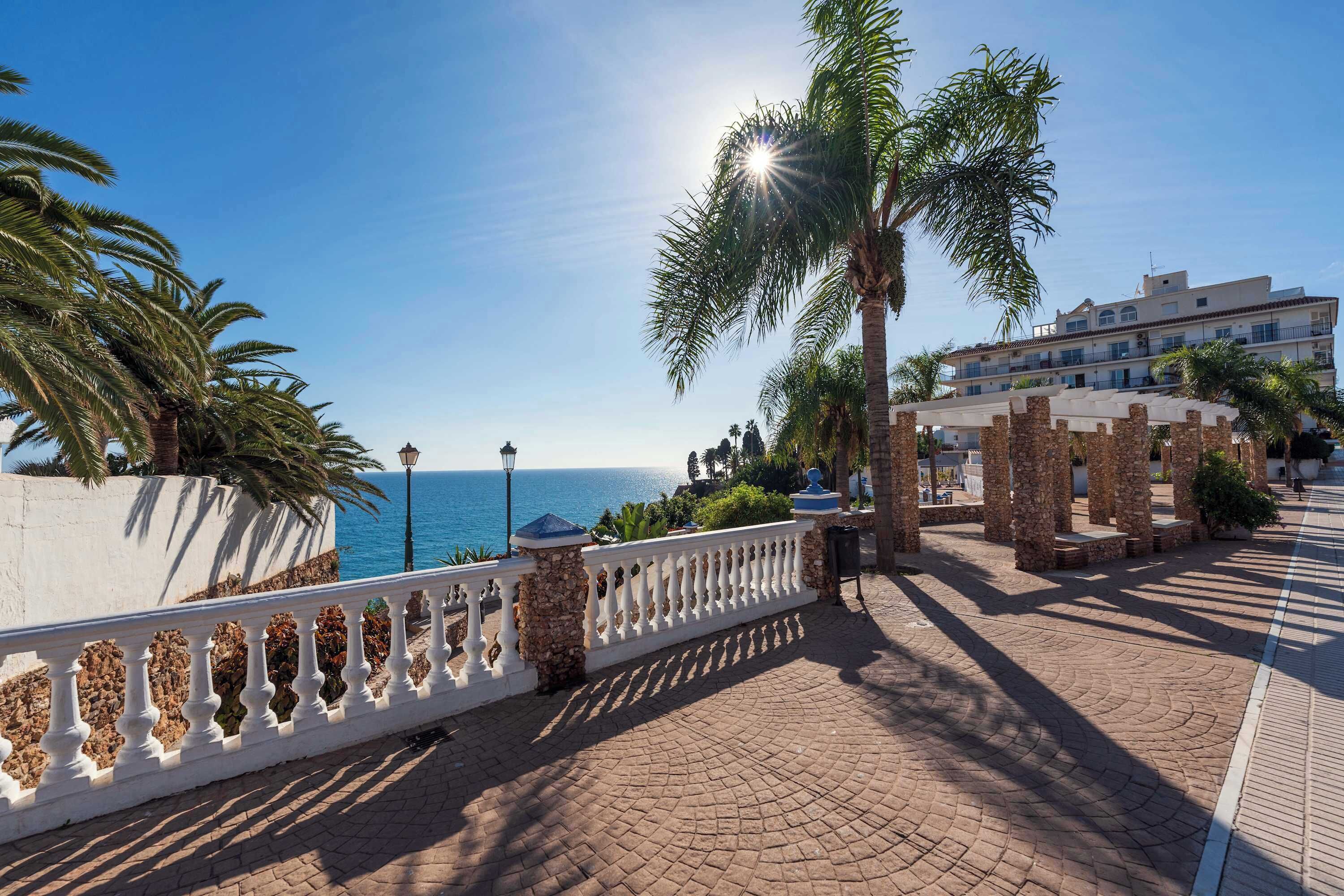 A coastal promenade with a white balustrade, palm trees, and a building with a tiled roof.