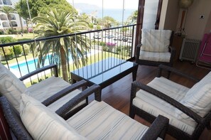 A balcony with wicker furniture, a view of a pool and greenery, and a glimpse of a building in the background. - Carabeo 1-30 (Nerja)