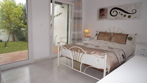 A bedroom with a bed, a bedside table with a lamp, a window with a view of a garden, and a wall with a piece of art. - Paraiso Blanco (Nerja)