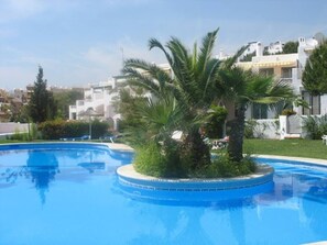 Une piscine avec un îlot circulaire, des palmiers et un bâtiment blanc en arrière-plan.
