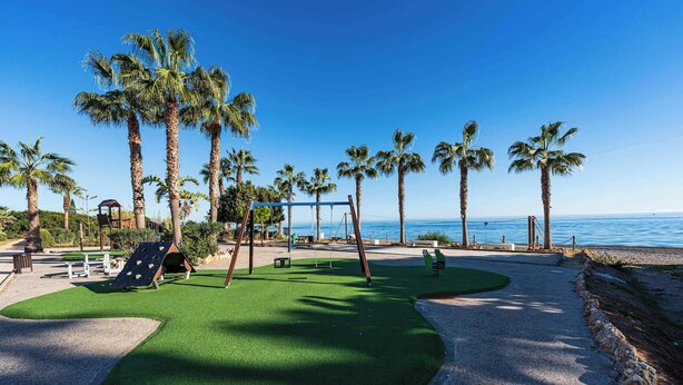 A playground with swings and slides, palm trees, and a view of the ocean.