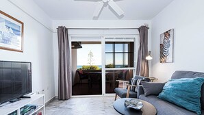 A living room with a sofa, a coffee table, a TV on a stand, and a bookshelf. There is a sliding glass door leading to an outdoor area with a view of the sea.