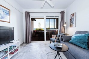 A living room with a sofa, a coffee table, a TV on a stand, and a bookshelf. There is a sliding glass door leading to an outdoor area with a view of the sea.