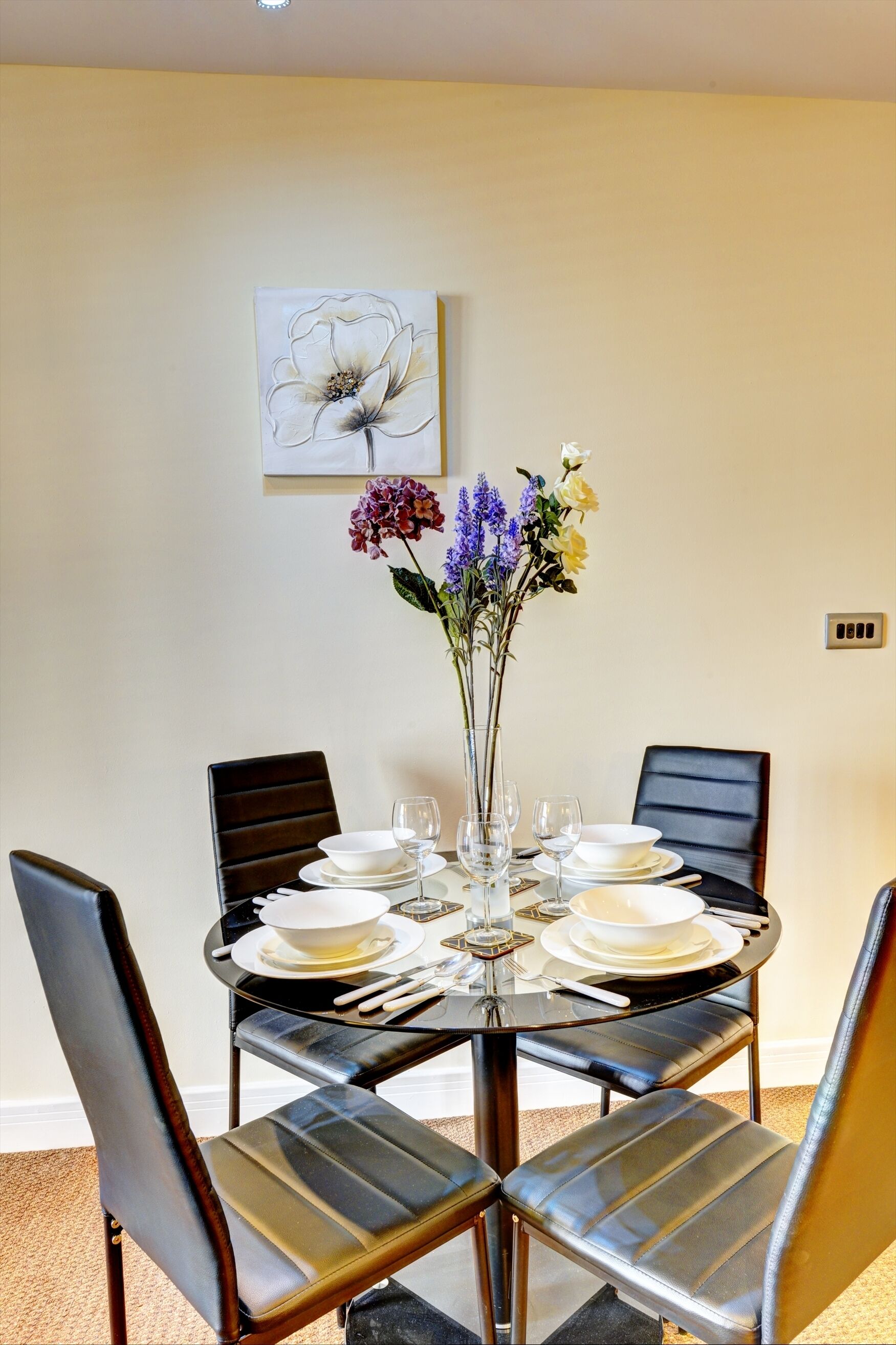 A dining table set for four with a vase of flowers, black chairs, and a painting on the wall.
