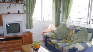 A living room with a sofa, a television, a table with fruit, and a window with curtains. - Edf Malaga (Nerja)