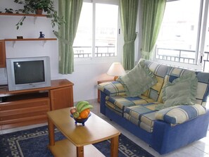 A living room with a sofa, a television, a table with fruit, and a window with curtains. - Edf Malaga (Nerja)