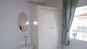 A bedroom with a white wardrobe, a bedside table with a vase, a mirror, and a window with curtains. - Edf Malaga (Nerja)
