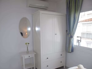 A bedroom with a white wardrobe, a bedside table with a vase, a mirror, and a window with curtains.