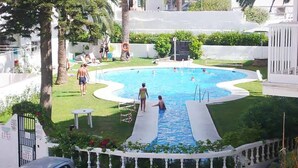 A swimming pool with a slide, surrounded by a white fence, palm trees, and a grassy area. - Edf Malaga (Nerja)