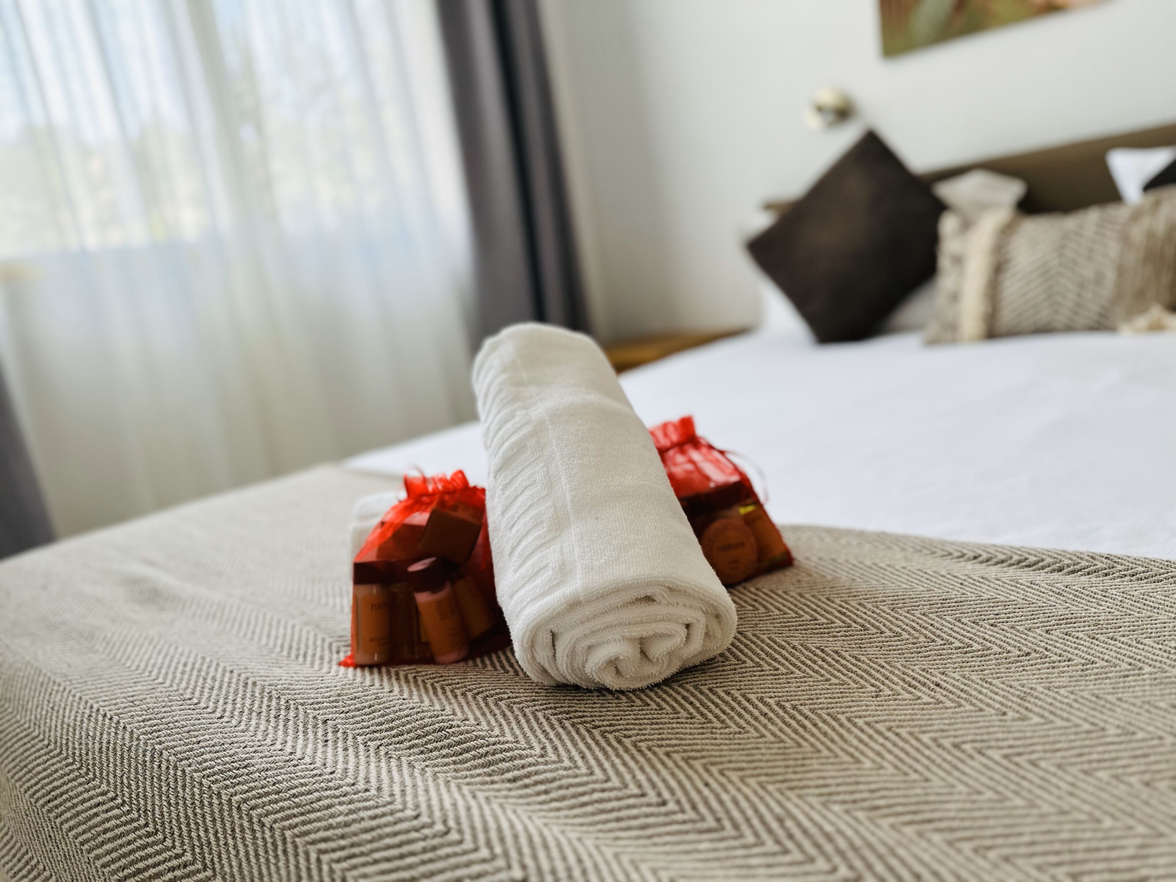 A neatly made bed with a rolled white towel and two red gift boxes on a textured beige bedspread.