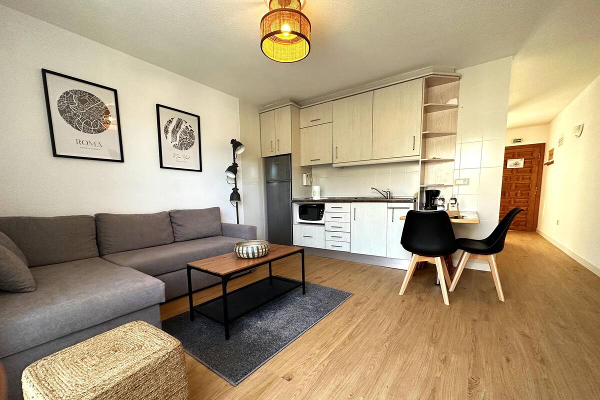 Un salon moderne avec un canapé gris, une table basse en bois et un fauteuil noir. On y trouve également un coin cuisine avec des meubles blancs et un plan de travail noir.