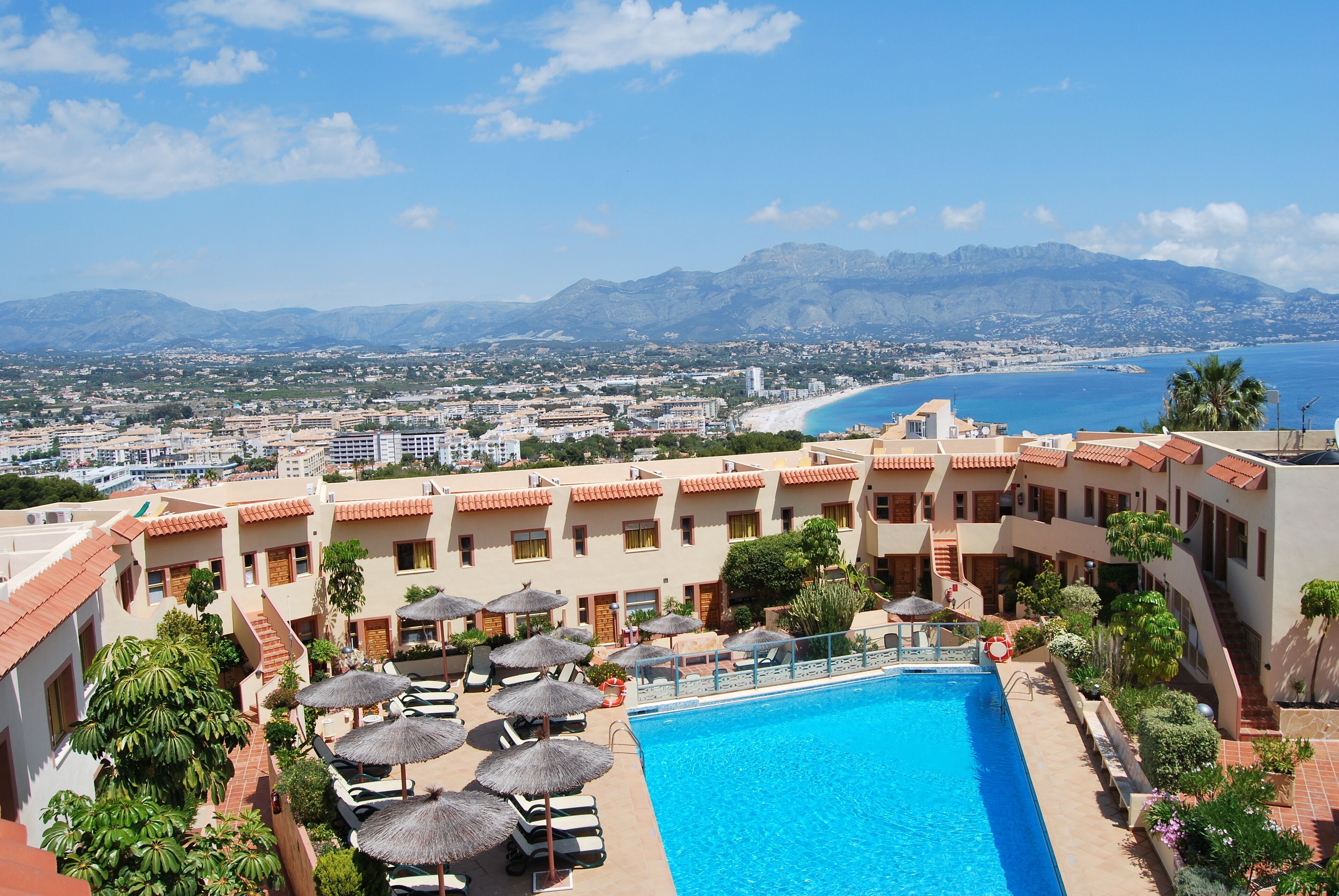 Ein Hotel mit Swimmingpool, Liegestühlen und Sonnenschirmen. Im Hintergrund Blick auf Strand und Berge.