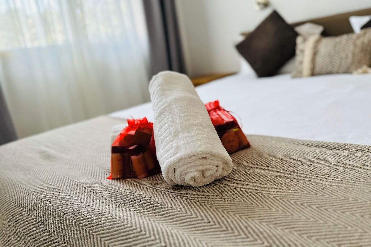 A neatly made bed with a rolled white towel and two red gift boxes on a textured beige bedspread.