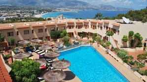 En resort-pool med udsigt til hav og bjerge, omgivet af liggestole og parasoller.