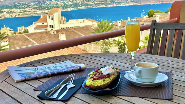 Et bord med en avis, en kop kaffe, et glas appelsinjuice og en tallerken med mad på en balkon med udsigt over en kystby og bjerge.