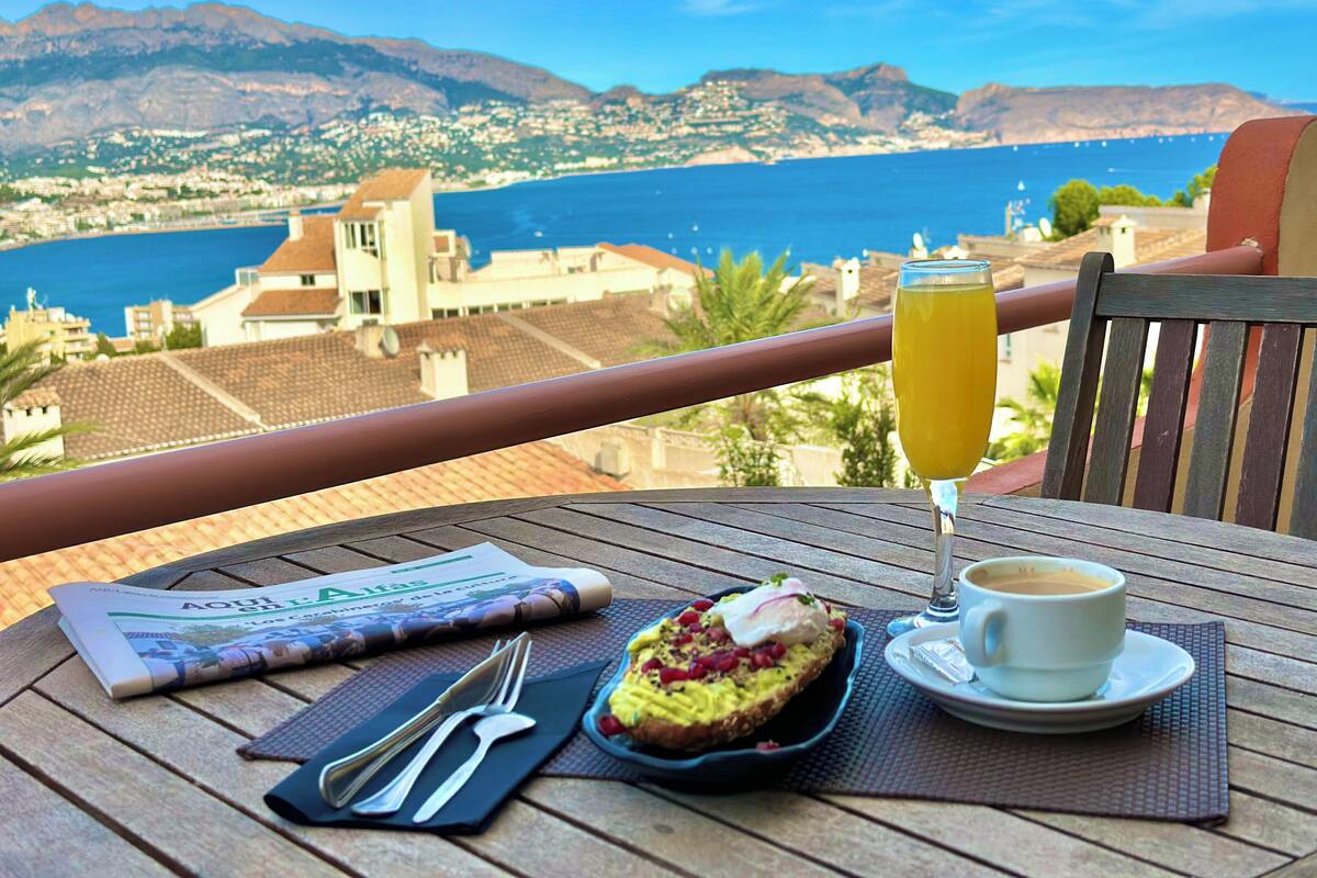 Une table avec un journal, une tasse de café, un verre de jus d’orange et une assiette de nourriture, le tout placé sur un balcon donnant sur une ville côtière et des montagnes.