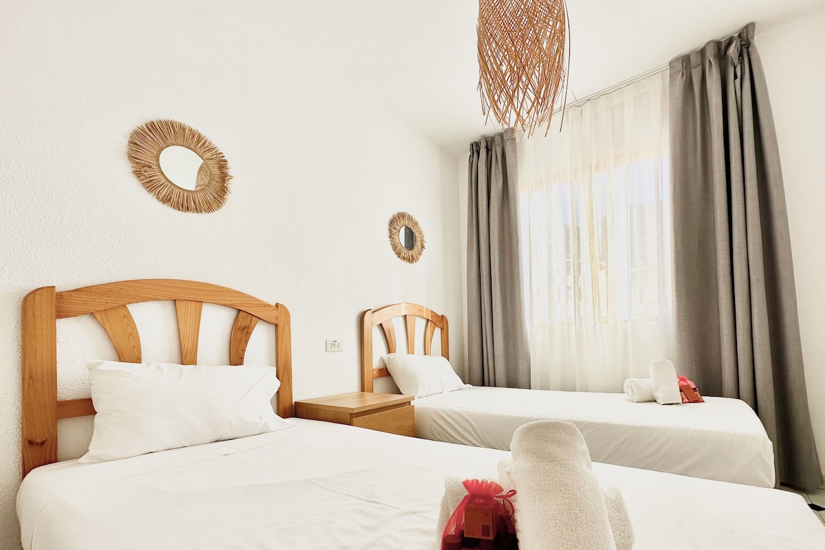 A hotel room with two beds, a wooden headboard, a small bedside table, a mirror, and a window with curtains.