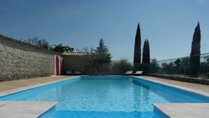 Une piscine rectangulaire à l’eau d’un bleu limpide, entourée d’un mur de pierre et bordée d’une terrasse carrelée. Deux chaises longues se trouvent sur le côté droit de la piscine.