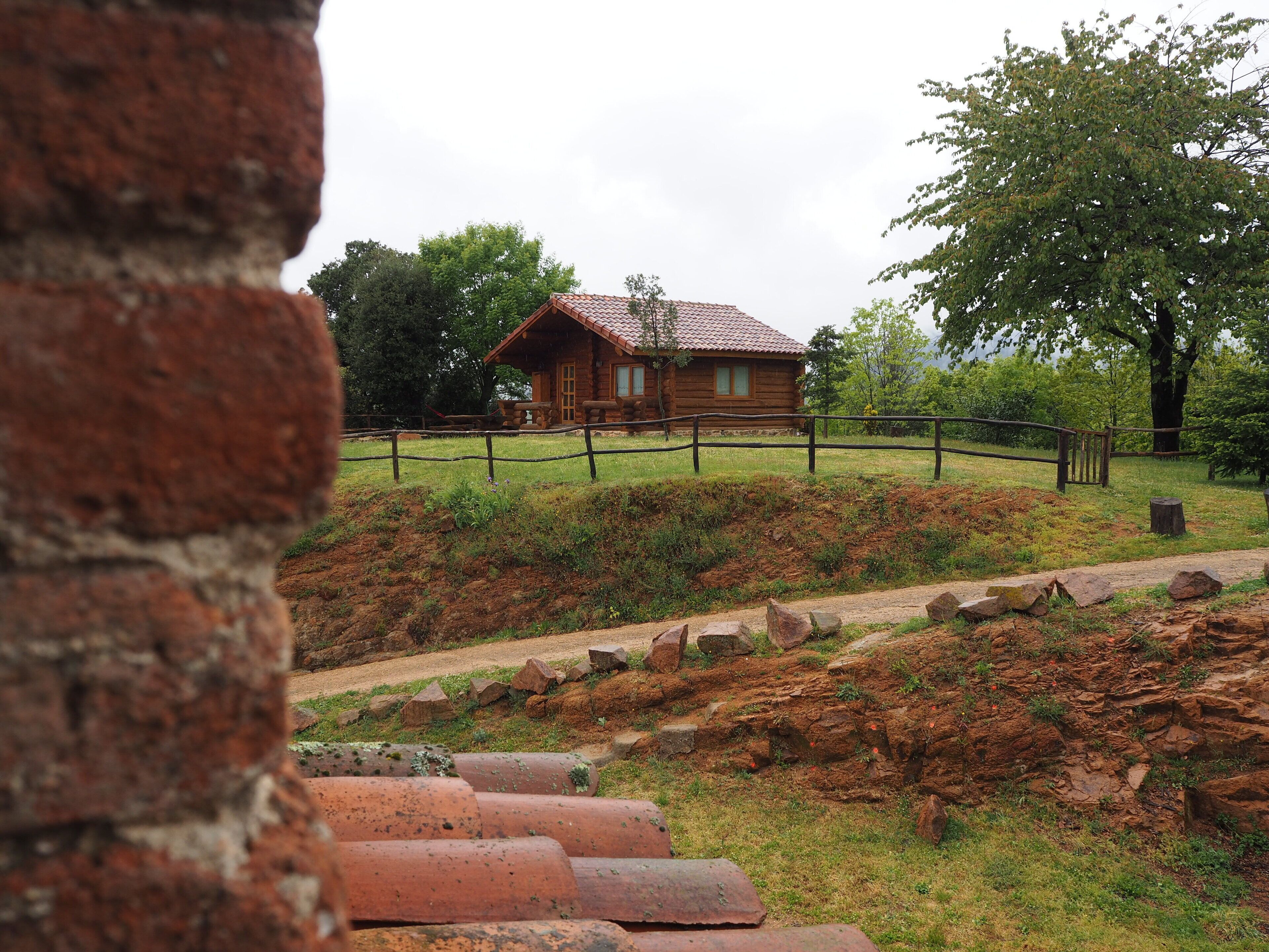 Una cabaña rústica de troncos con un patio cercado, rodeada de árboles y un sendero de tierra que conduce a ella.