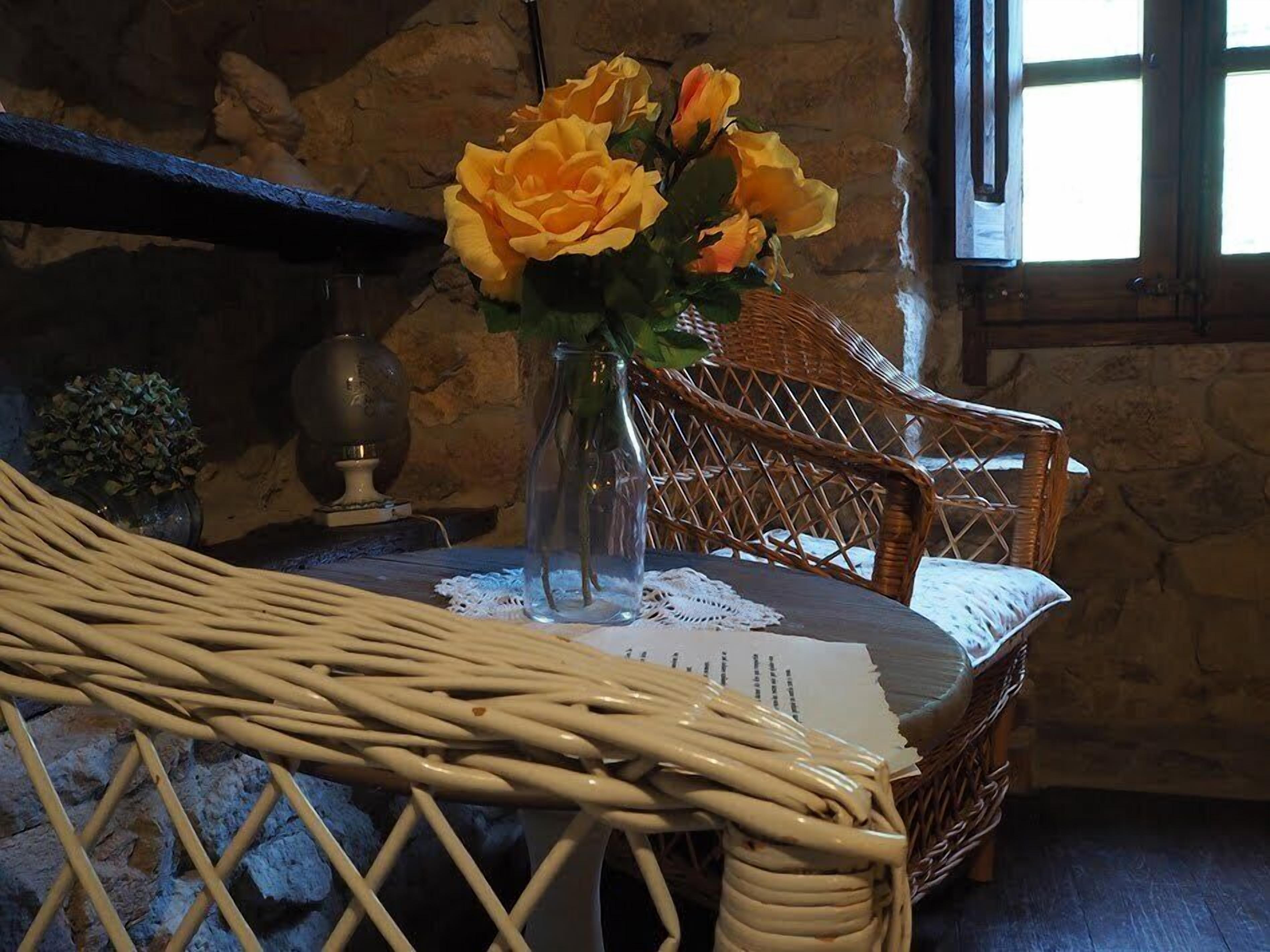 Une chaise en osier ornée d’un chemin de table en dentelle, un vase avec des roses jaunes et, en arrière-plan, un mur de pierres.