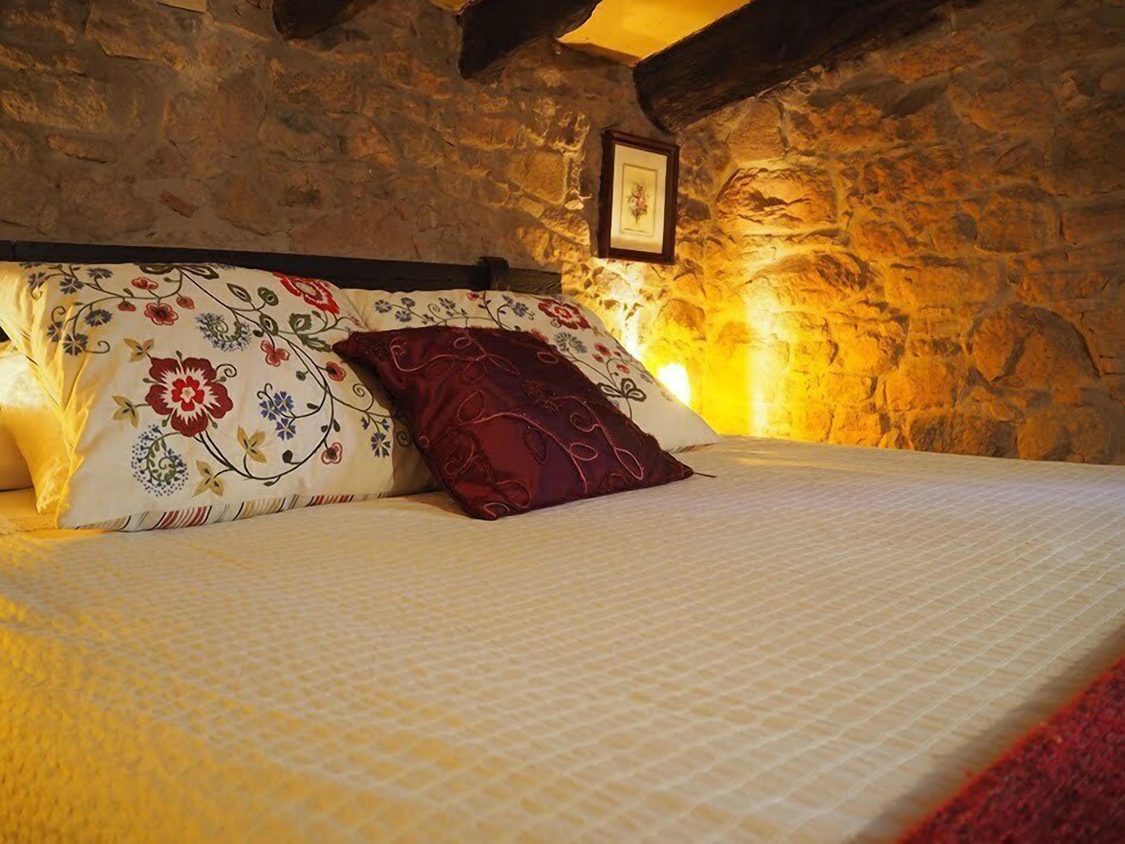 A bed with floral-patterned pillows and a red accent pillow in a room with stone walls and exposed wooden beams.