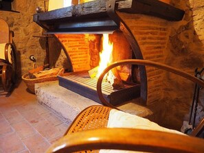 A stone fireplace with a fire burning inside, a wooden bench, and a woven basket on the floor. - Mas Cal Sastre (Sant Hilari Sacalm)