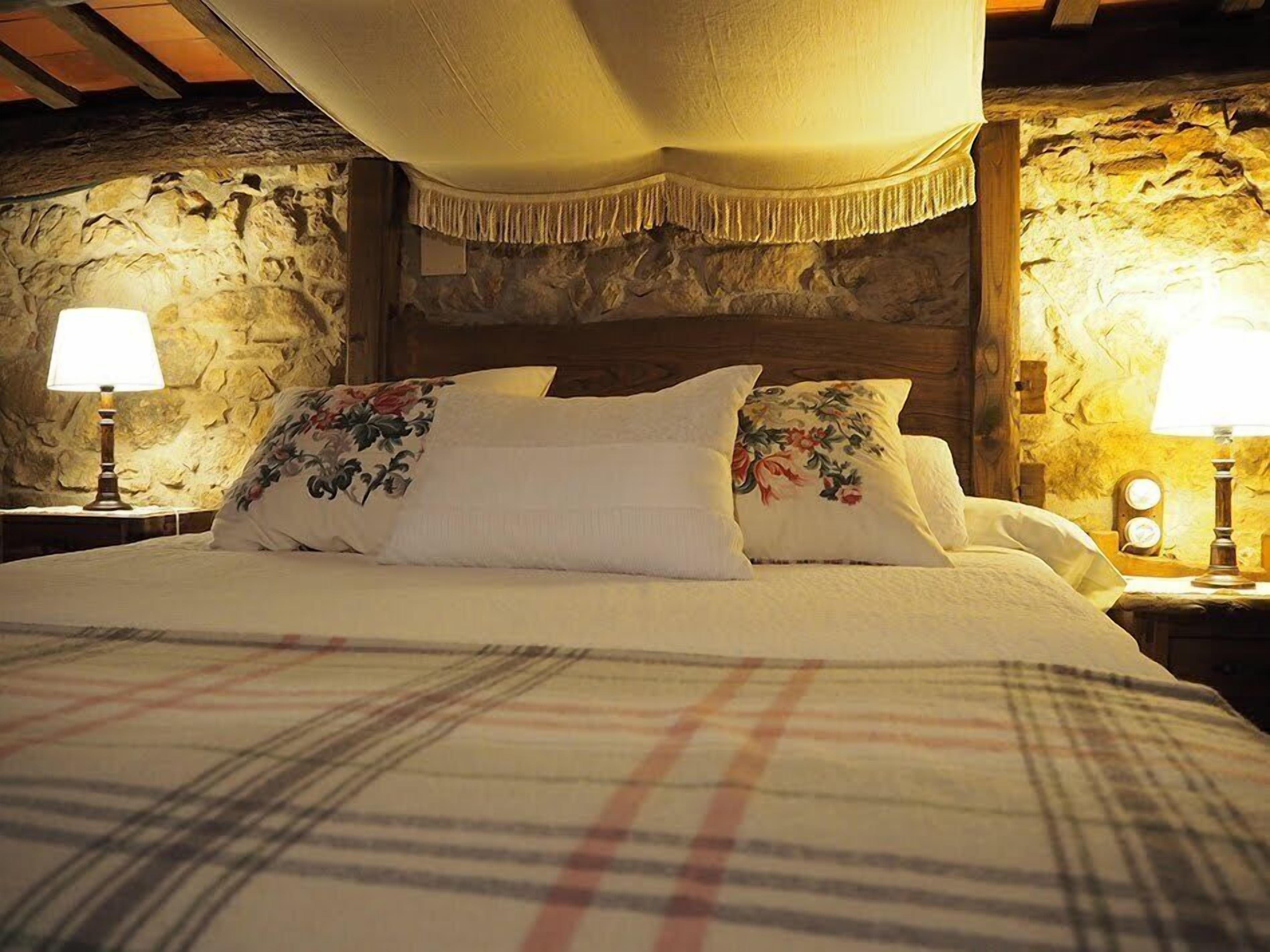 A bed with floral pillows, a plaid blanket, and a stone wall backdrop.