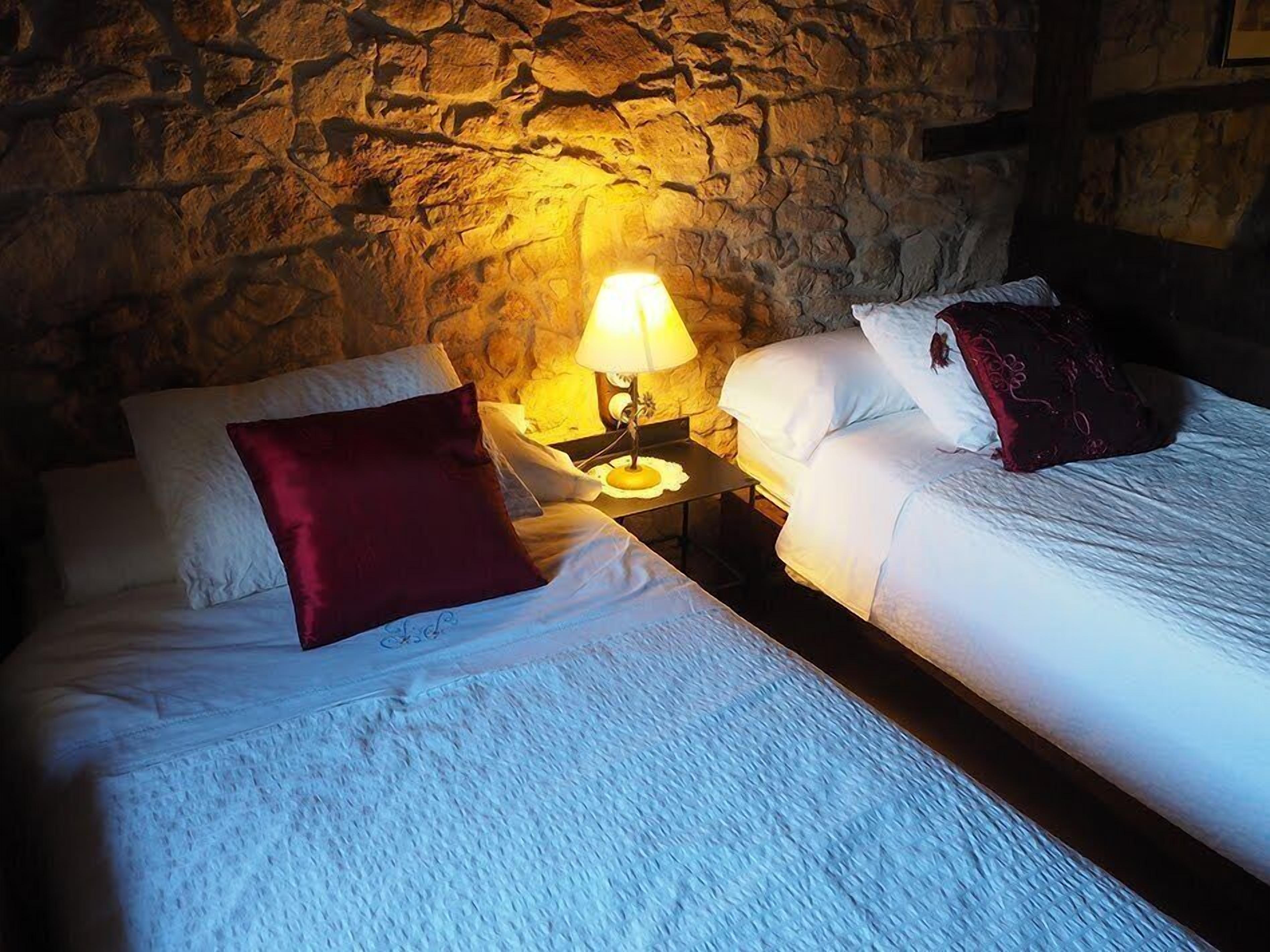 Una habitación con dos camas, cada una con ropa blanca y una almohada roja. Hay una pared de piedra detrás de las camas y una lámpara en una mesita de noche.