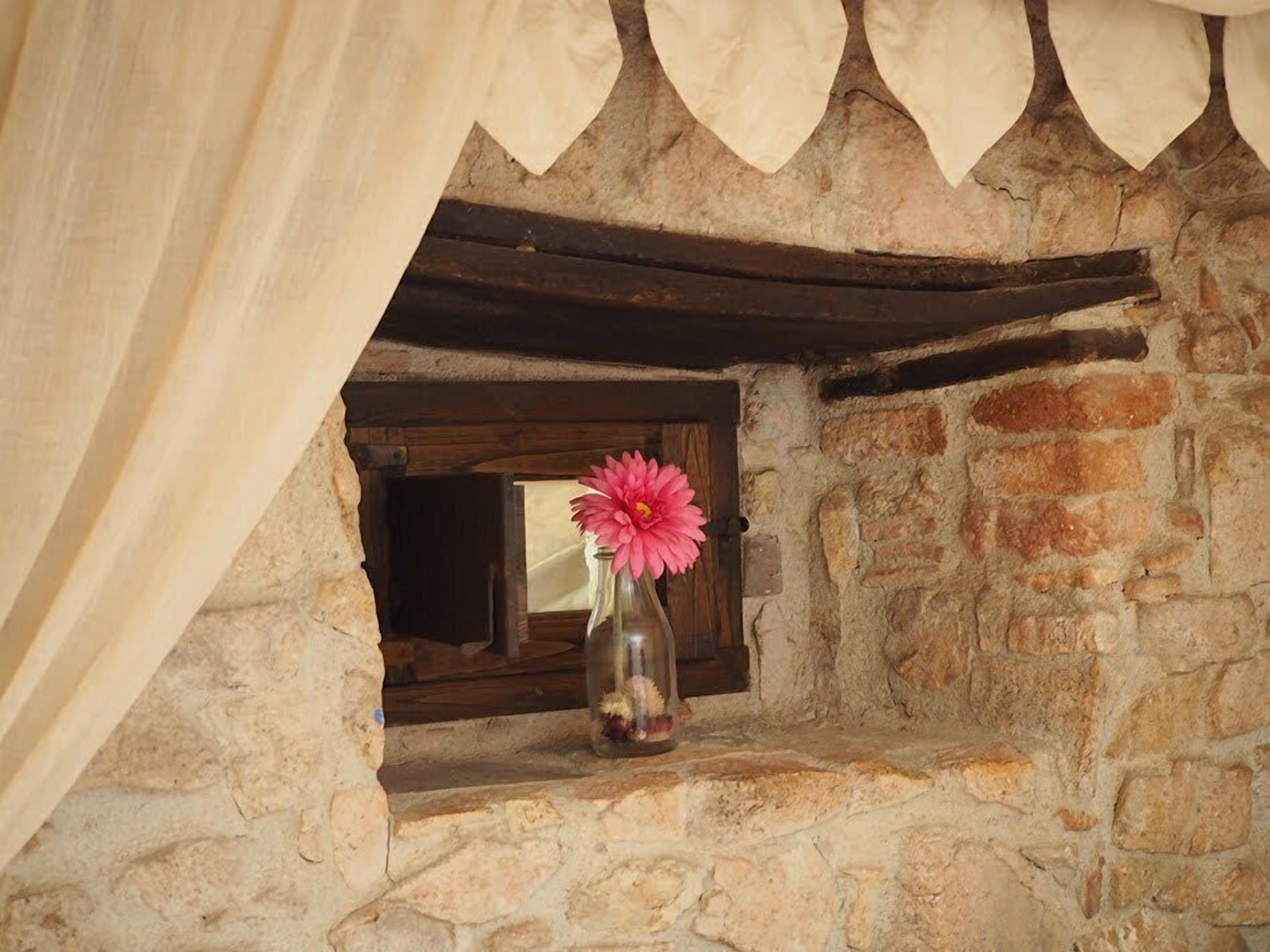 A stone fireplace with a window, a vase with a flower, and a curtain.