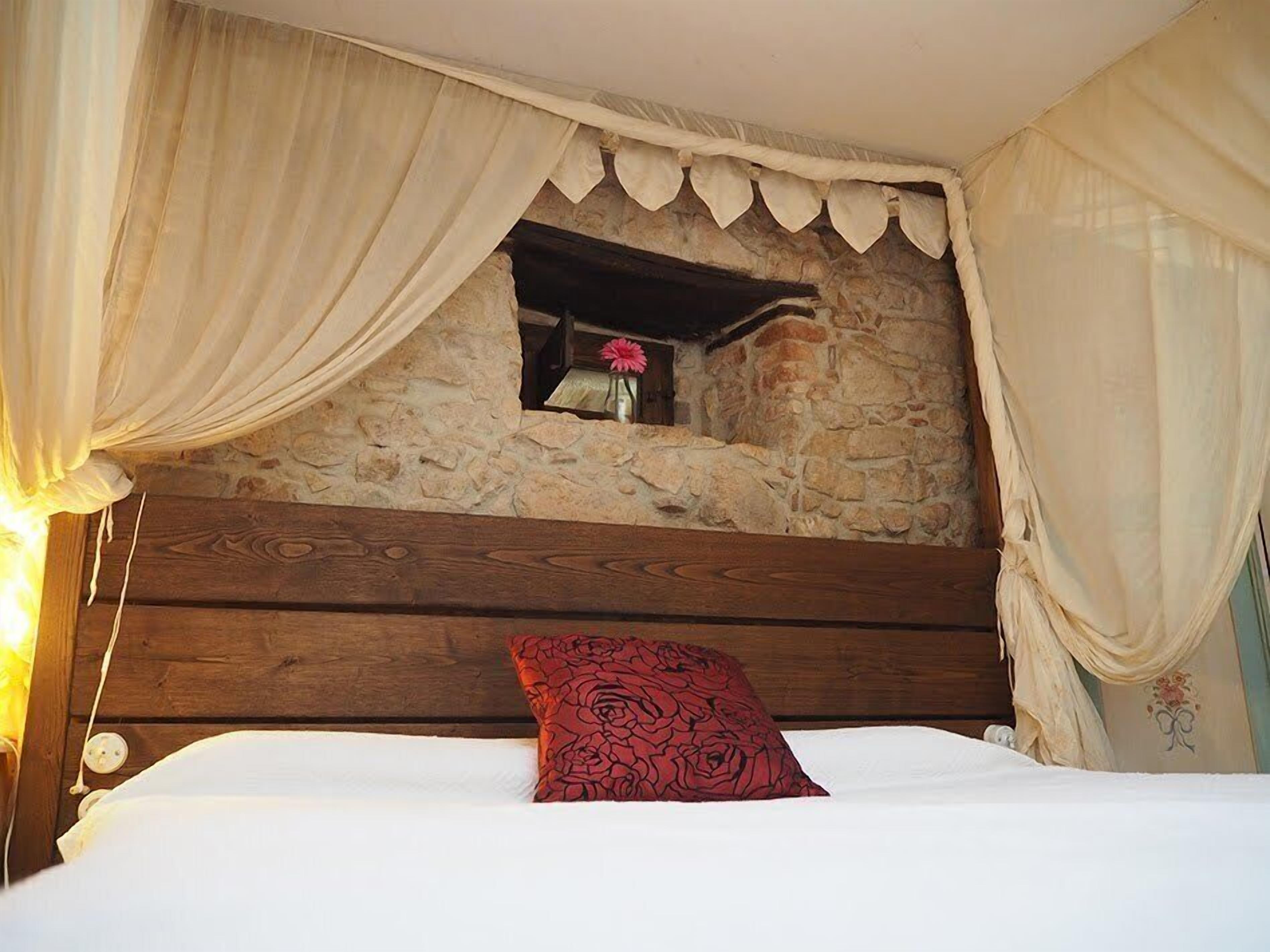 A bedroom with a stone wall, wooden headboard, and a window with a flower vase.
