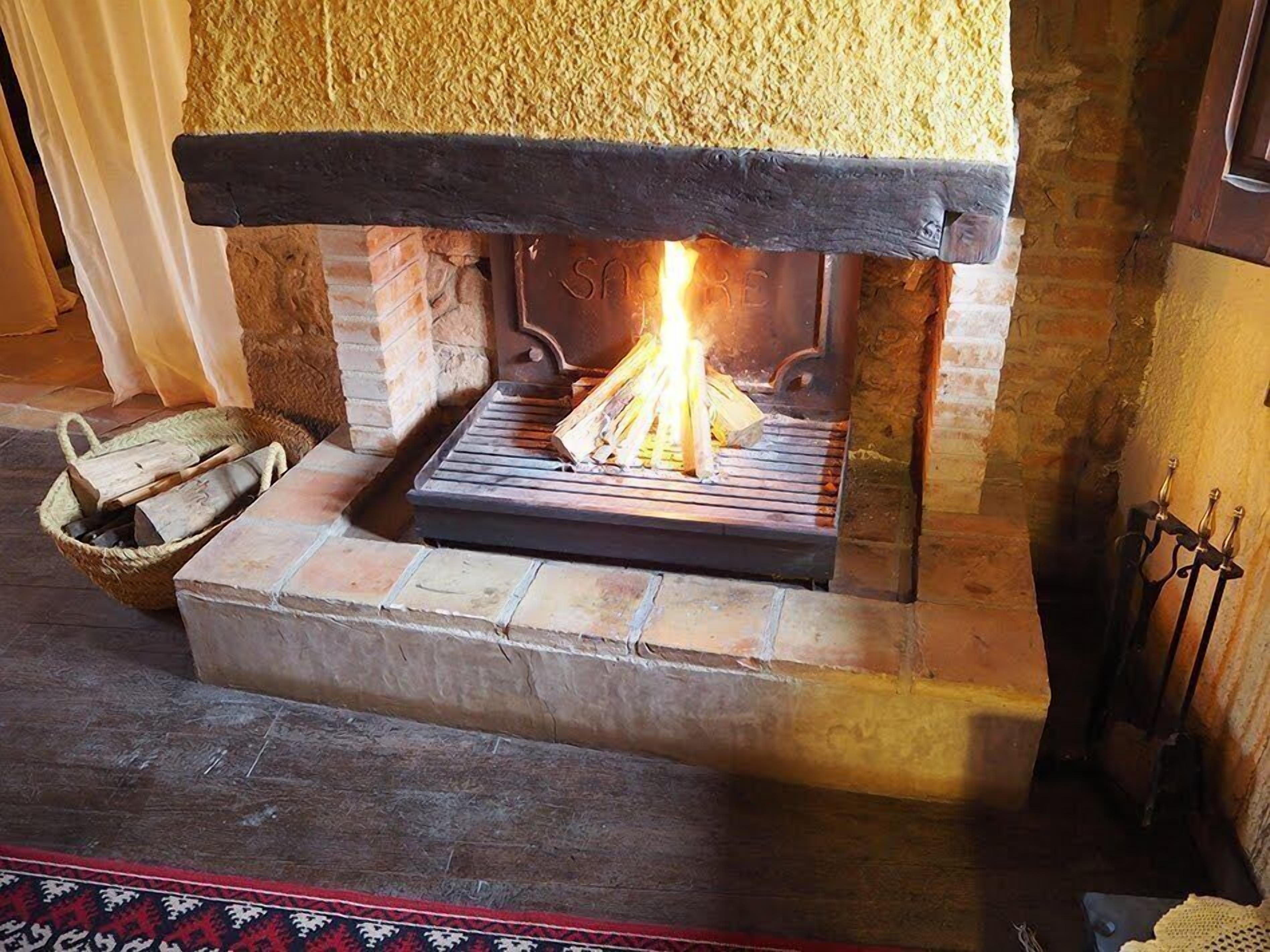 A stone fireplace with a fire burning inside, a basket of firewood, and a metal grate.
